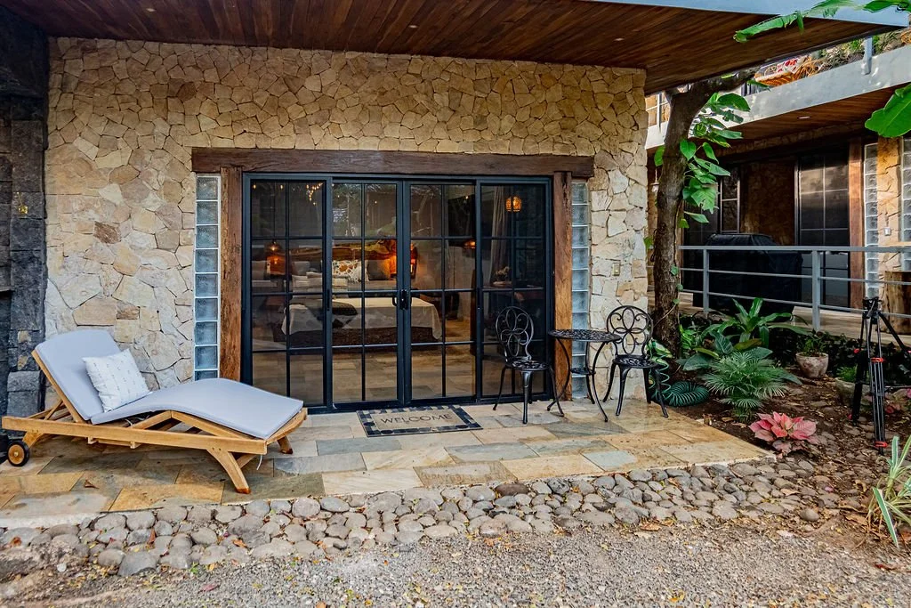 A cozy outdoor patio area in front of a stone house with sliding glass doors, featuring a lounge chair with a cushion, a small round table, two black metal chairs, and lush green plants.