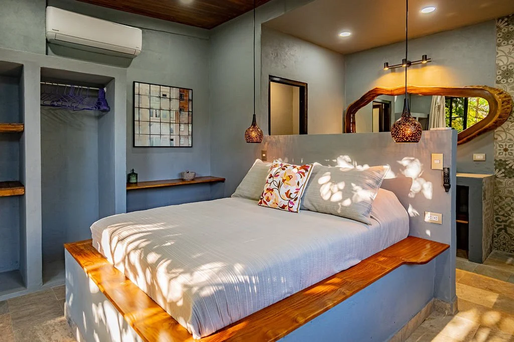 A cozy bedroom with a bed featuring white bedding, decorative pillows, and a rustic wooden headboard. Hanging pendant lights illuminate the space, and a large mirror reflects the room's warm lighting and wooden accents. There is an air conditioning u