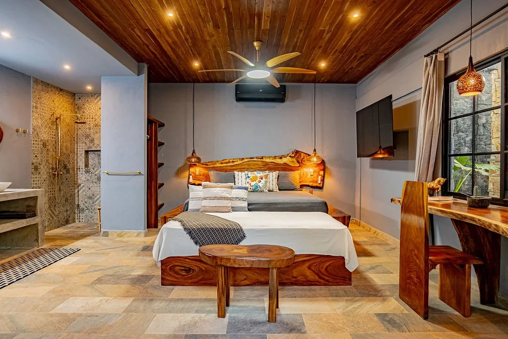 A cozy modern bedroom with a wooden ceiling, king-sized bed, and decorative pillows. There is a small wooden bench at the foot of the bed, a flat-screen TV mounted on the wall, and a large window with beige curtains. An open bathroom area with a show