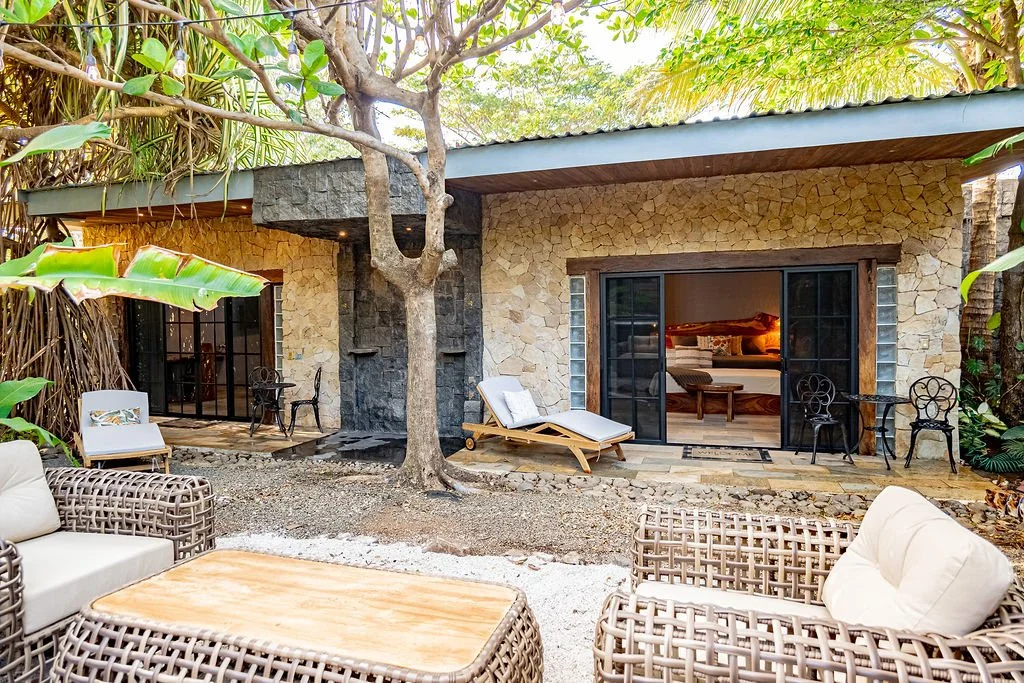 Outdoor patio of a house with stone walls, glass sliding doors, and tropical plants, featuring lounge chairs, wicker furniture, and a tree in the center.