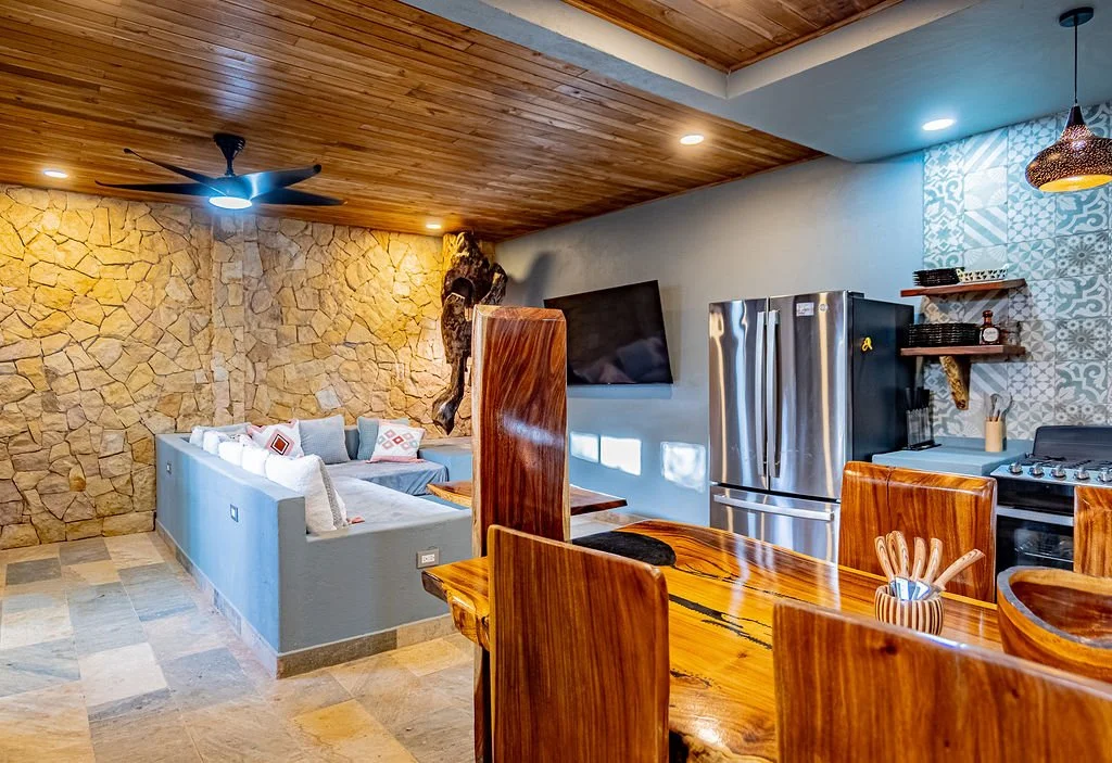 Open-concept living room and kitchen with stone accent wall, wooden ceiling, and contemporary furniture.