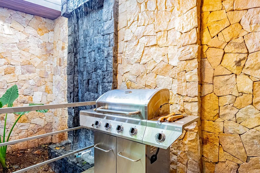 Stainless steel barbecue grill with four control knobs, placed against a stone wall with a black water feature wall to the left.