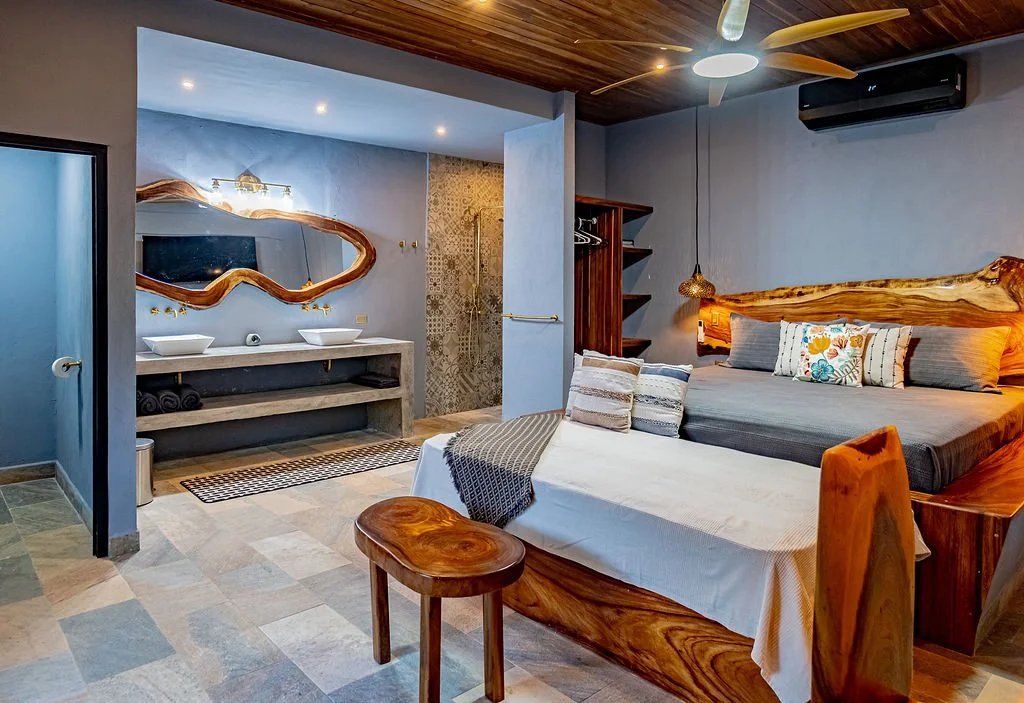 A hotel room with a wooden bed, multiple pillows, a mounted TV, a granite-topped double sink vanity with a decorative mirror, open closet shelves, a ceiling fan, and a bathroom area with a tiled shower.