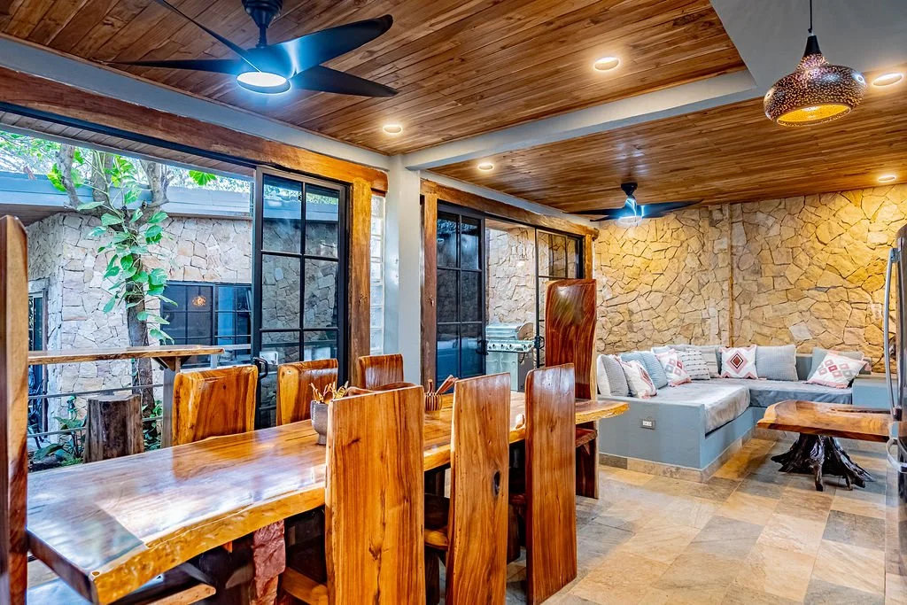 A cozy living room and dining area with wooden furniture, stone accent walls, and large glass doors leading to an outdoor space with trees.
