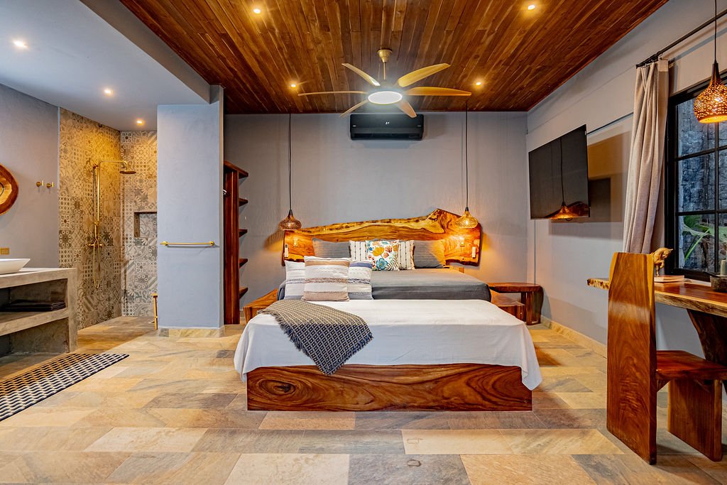 A cozy bedroom with a wooden bed and headboard, a large window with curtains, a wall-mounted TV, and an en-suite bathroom with a walk-in shower visible in the background.