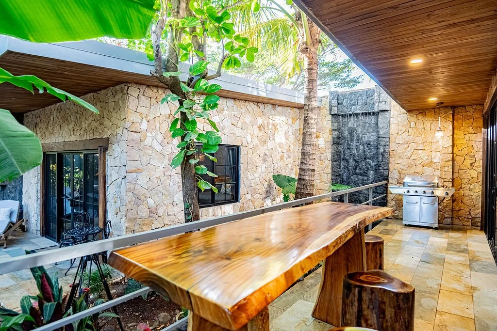 Outdoor patio with a polished wood bar table and stools, a stone wall with a small waterfall feature, lush tropical plants, and a stainless steel grill on a tiled floor, surrounded by tropical trees and greenery.
