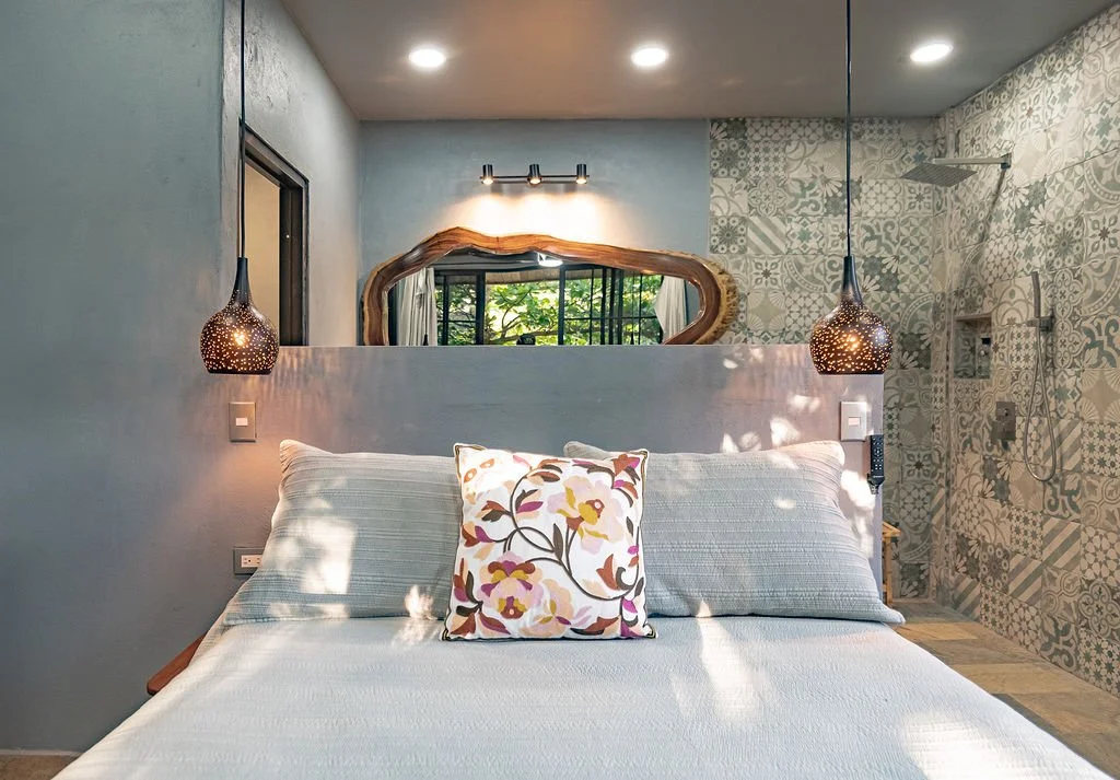Interior of a bedroom with a bed, decorative tufted pillow, hanging pendant lamps, mirror, and patterned shower area in the background.