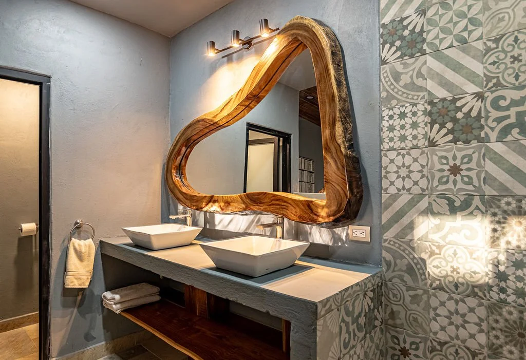 Modern bathroom with two vessel sinks on a white countertop, a large irregularly shaped wooden framed mirror, geometric patterned tiles on one wall, gray walls, a wall-mounted light fixture, and folded towels on a wooden shelf.