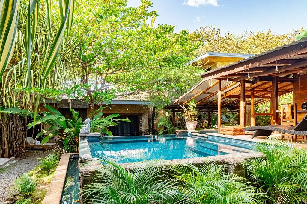 Tropical backyard with a small pool surrounded by lush green plants and trees, a covered patio area with wooden posts, a lounge chair, and decorative statues.