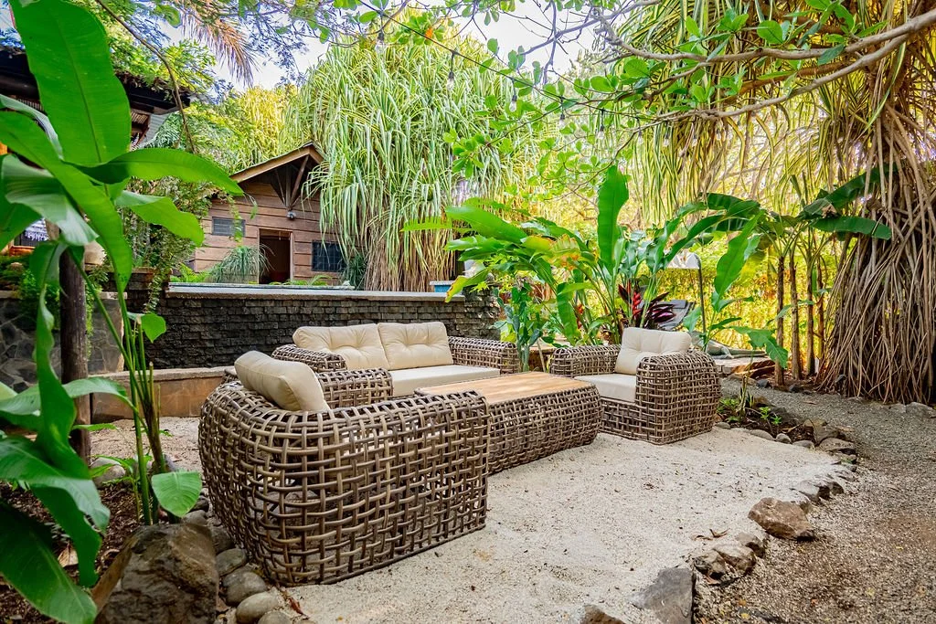 Outdoor seating area with wicker furniture and cushions, surrounded by lush green tropical plants and trees.