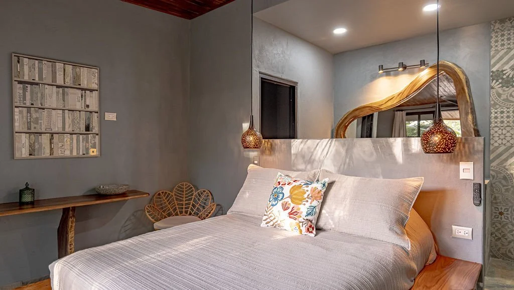 A bedroom with a bed, decorative pillows, a mirror above the headboard, hanging pendant lights, and a wooden console with a rattan chair.