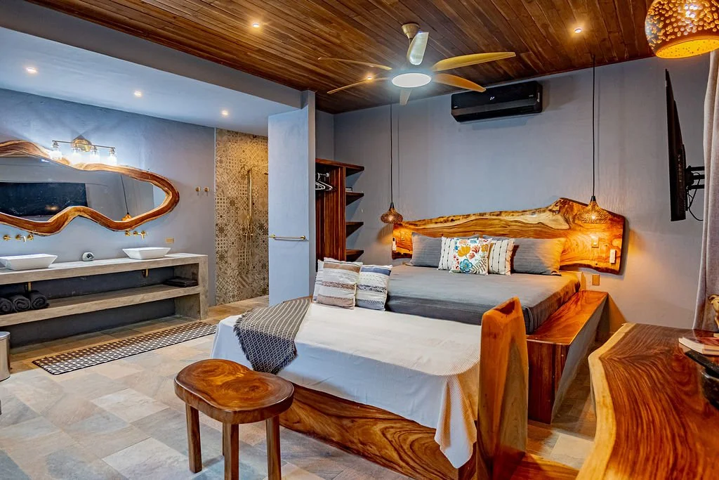 Modern bedroom with wooden bed frame, colorful pillows, and a beige bedspread. There is a wooden ceiling fan, a mounted TV, and a split air conditioner. A small wooden stool is beside the bed, and a bathroom area with sinks and a mirror is visible in