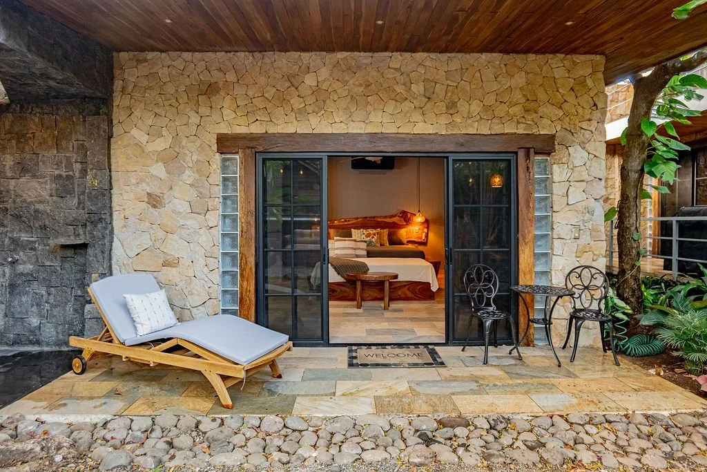 View of a cozy outdoor patio with a stone wall, wooden ceiling, and sliding glass doors leading into a bedroom with a bed and warm lighting. The patio has a lounge chair with a pillow, a small table with two chairs, and surrounding plants.