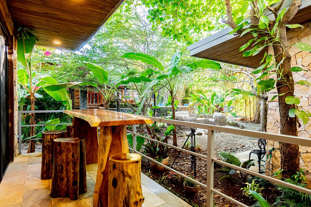 A cozy outdoor patio with a wooden bar table and matching stools, surrounded by lush green plants and trees, with patio chairs and a tripod in the background.