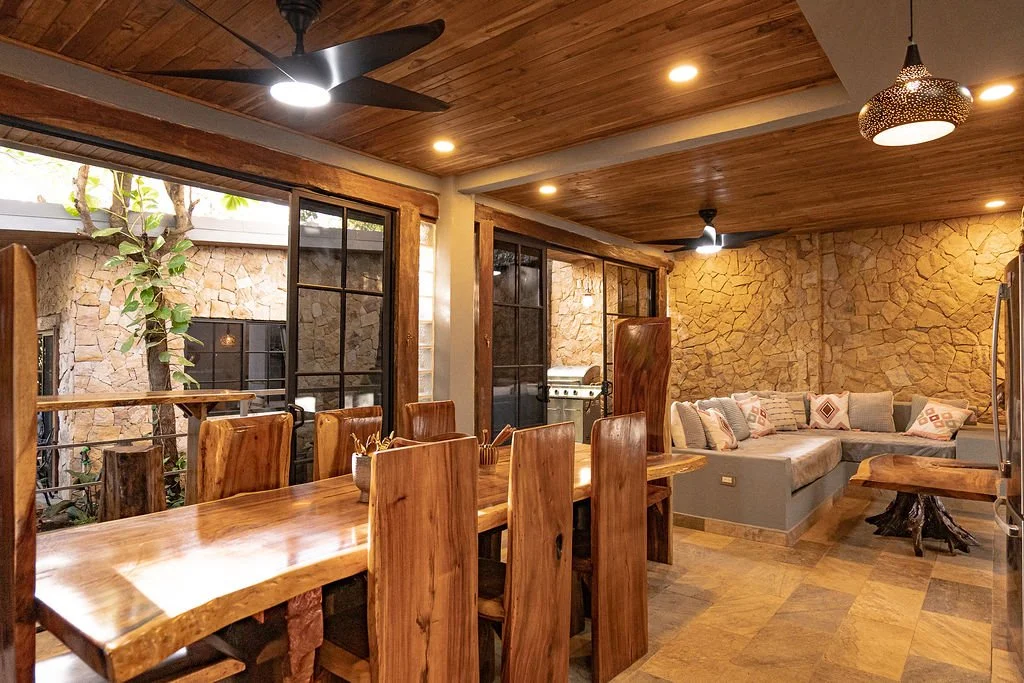 Cozy indoor living and dining area with stone walls, wooden ceiling, large window, wooden dining table with chairs, and a sectional sofa with cushions.