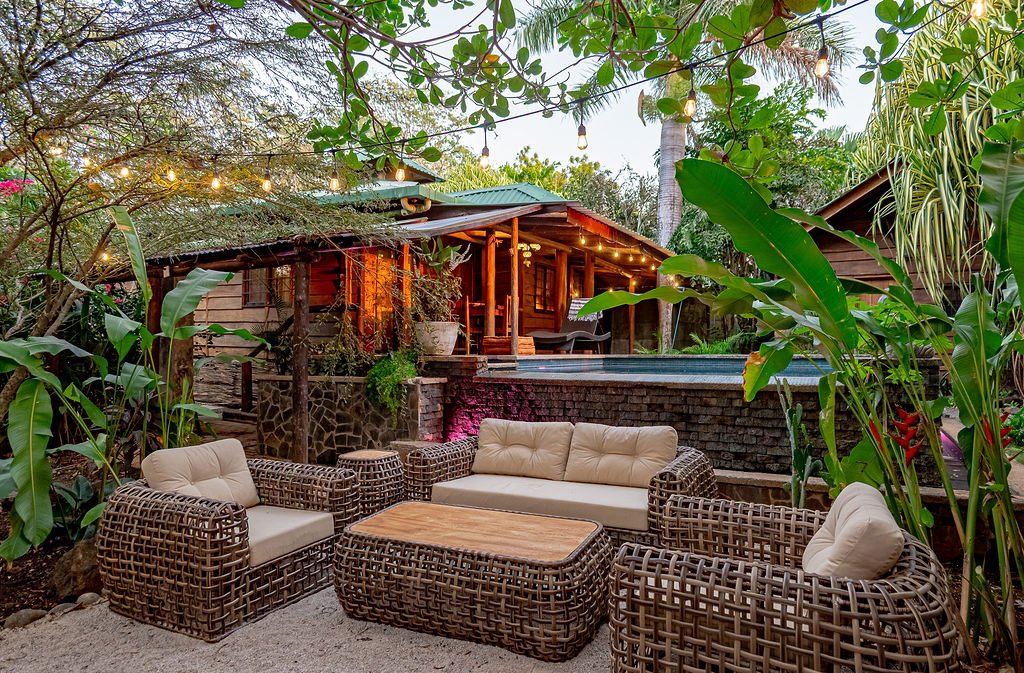 A cozy outdoor seating area with wicker chairs and a sofa, surrounded by lush green tropical plants, overlooking a backyard with a raised deck and a pool, illuminated with string lights at dusk.