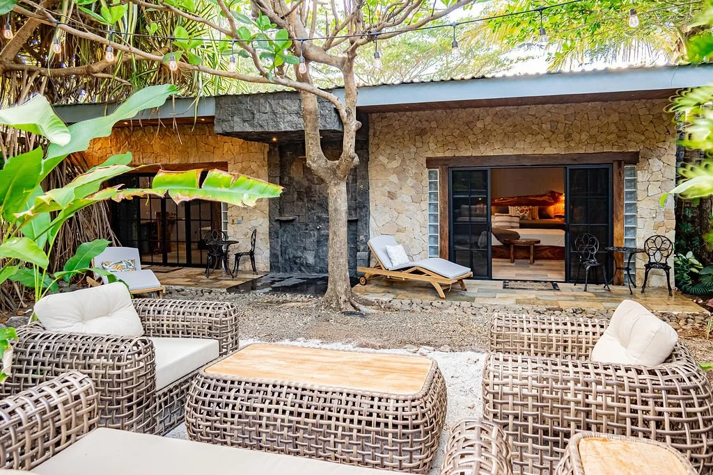 Outdoor patio area with wicker chairs and beige cushions, a wooden coffee table, a lounge chair, and lush green plants and trees surrounding a stone house with large sliding glass doors.