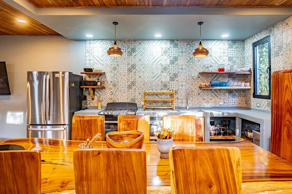 A cozy kitchen with a wooden dining table and chairs, stainless steel refrigerator, patterned tile backsplash, open shelves with dishes, and two hanging pendant lights.
