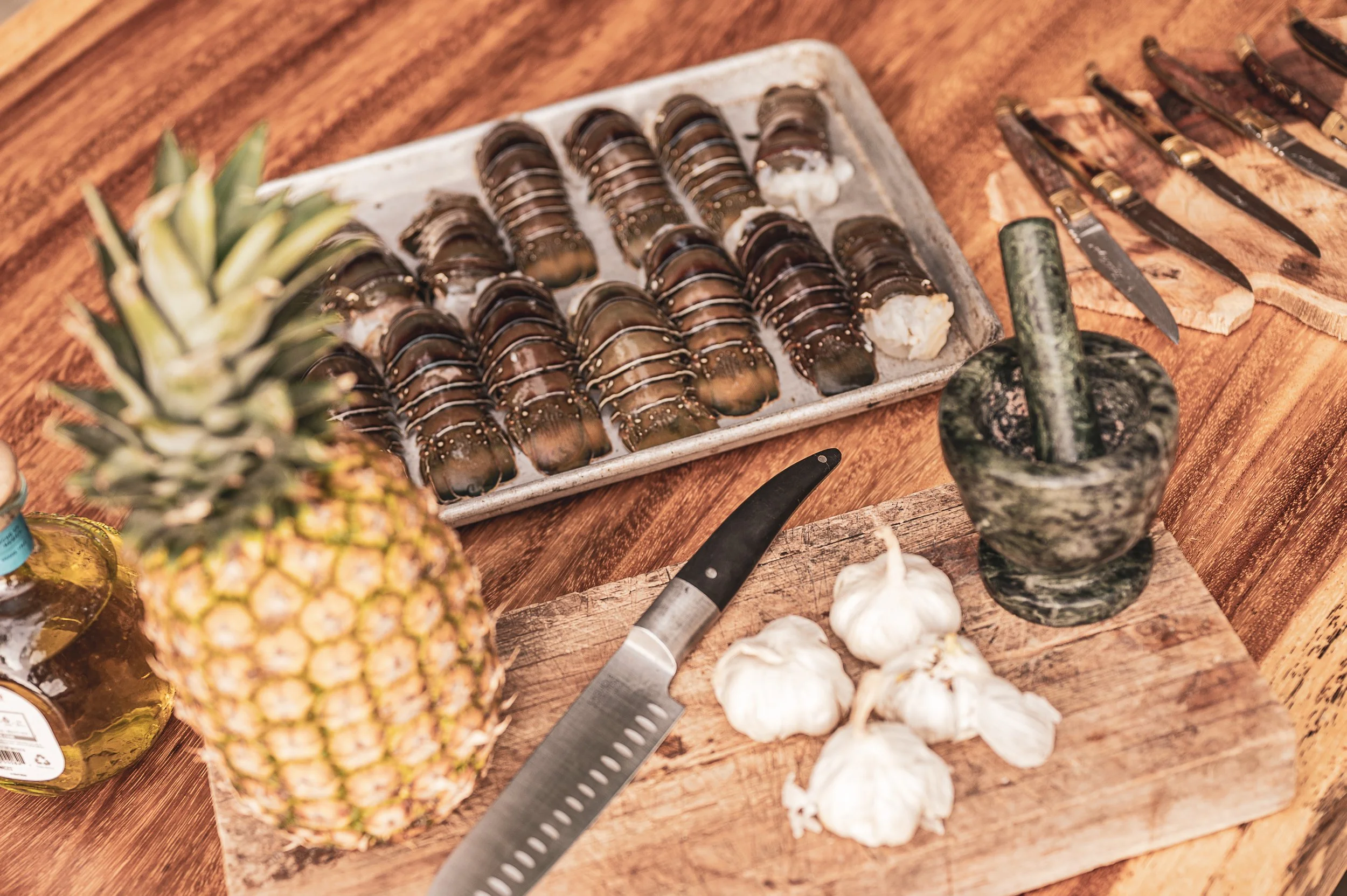 Tray of freshly prepared chocolate-covered éclairs on a rustic wooden table, with a pineapple in the foreground, garlic bulbs, a mortar and pestle, a chef's knife, and various knives in the background.