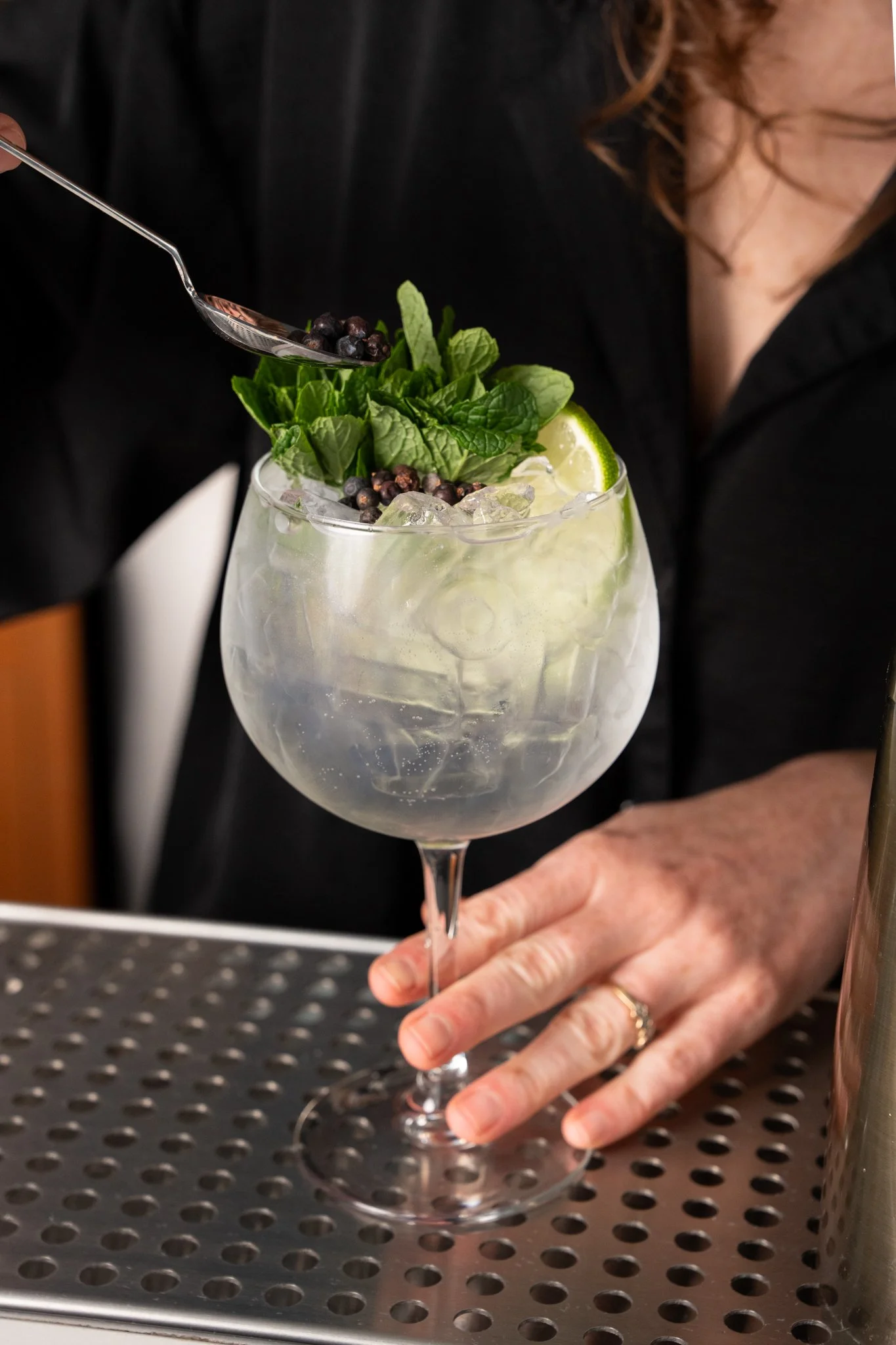 Bartender making Aria Gin Tonic Cocktail made with local gin, elderflower tonic, mint, lime, spooning white pepper on to large cocktail glass..