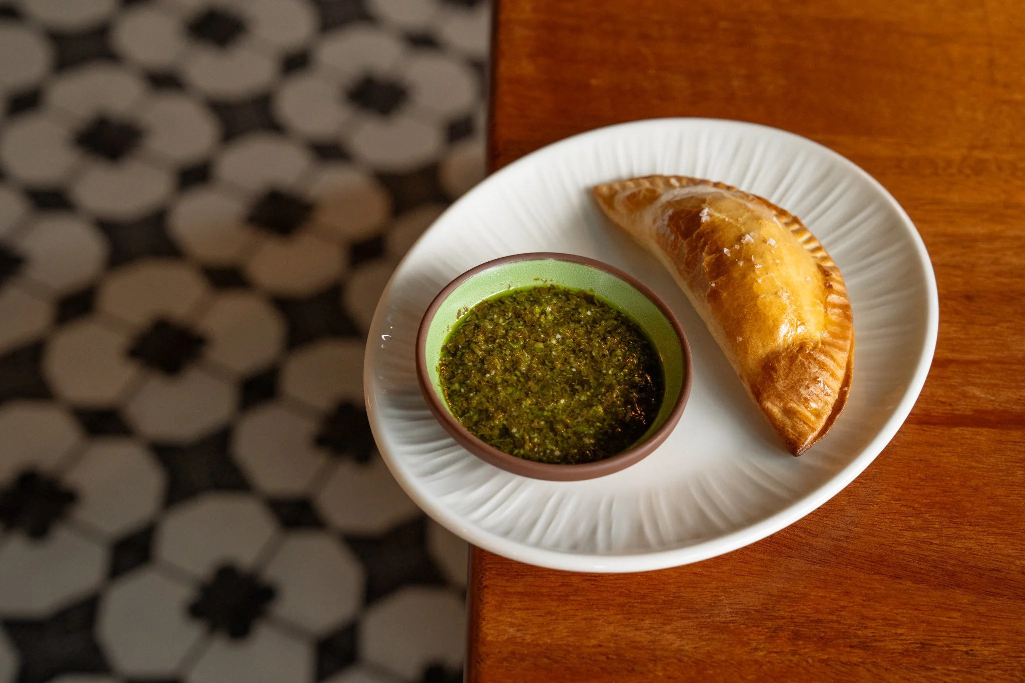 Oxtail empanada made with red wine braised oxtail, served with mojo verde, plated on a white plate sitting on a brown table.