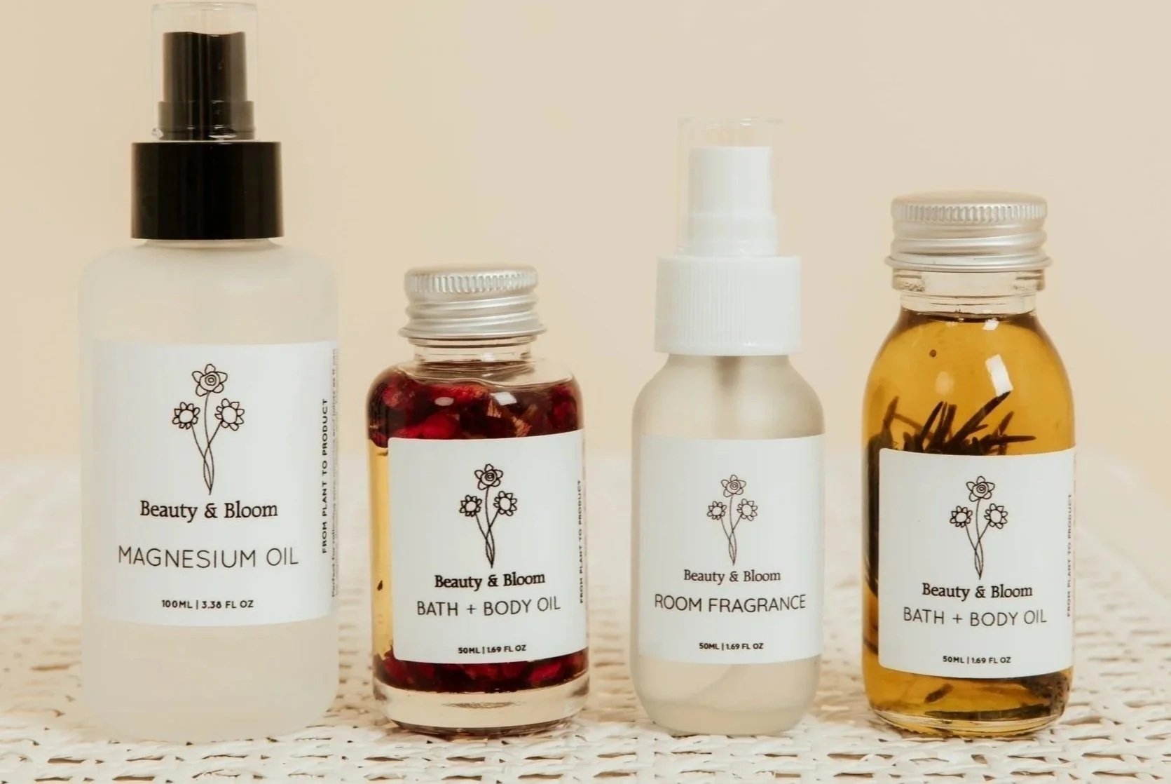 Set of four Beauty & Bloom personal care products including magnesium oil, bath and body oil, room fragrance, and another bath and body oil with herbal infusions, in clear glass bottles with white labels on a woven surface.