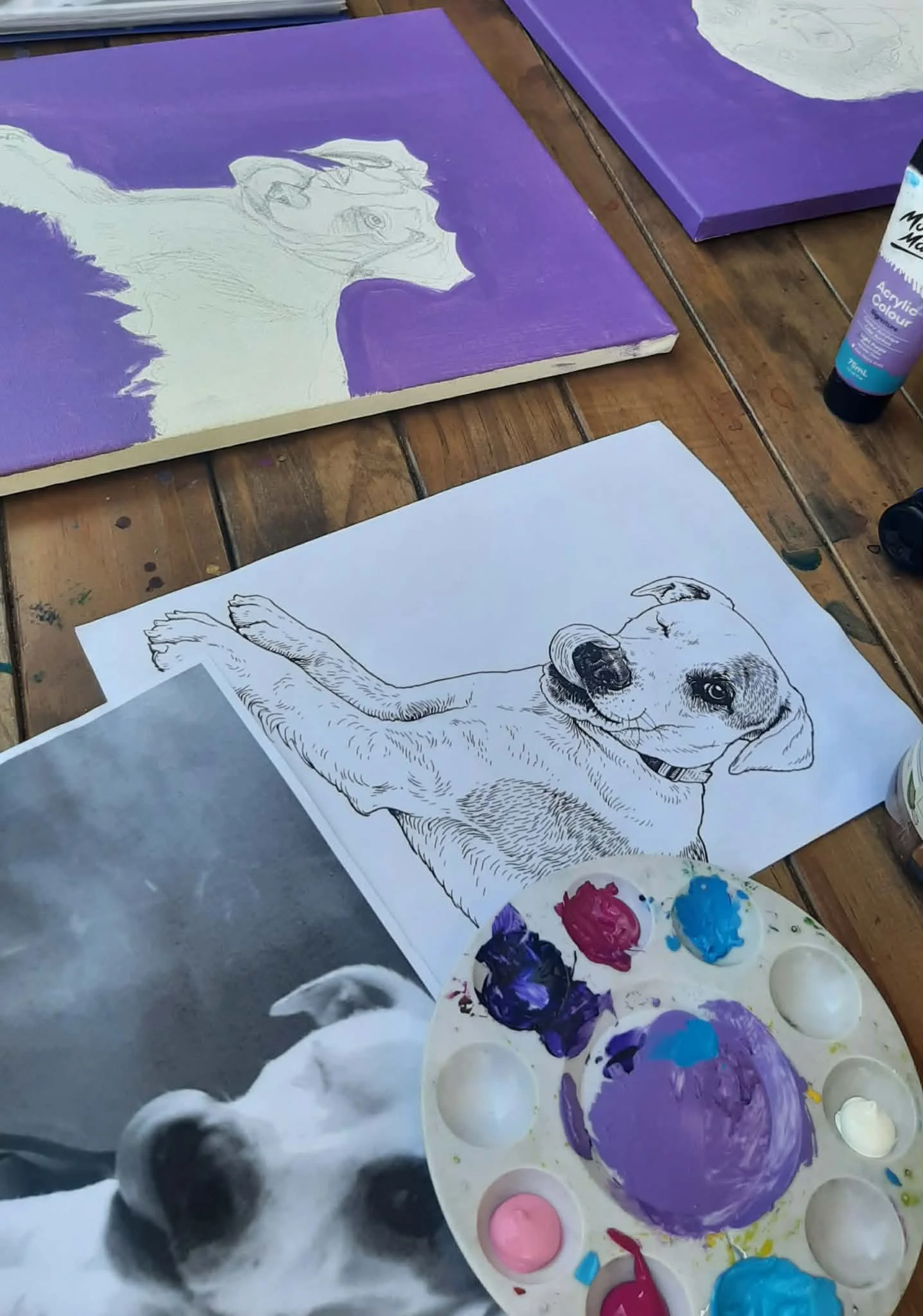 A table with a partially completed sketch of a dog, a printed photo of a dog, a palette with mixed paint, and an underpainting of a dog on a canvas.