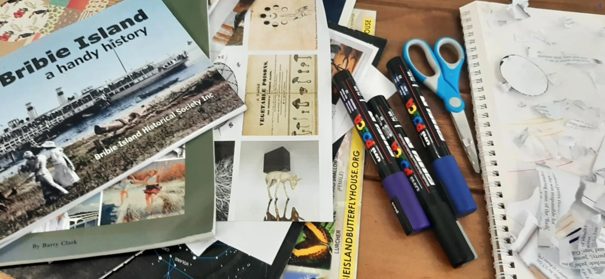 Watercolor painting supplies including watercolor paints, brushes, a completed watercolor with purple flowers, a pair of green-tinted sunglasses, and pink flowers on a wooden surface.