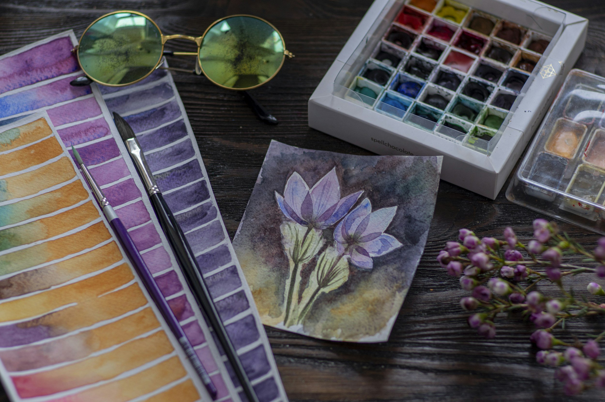 Watercolor painting supplies including watercolor paints, brushes, a completed watercolor with purple flowers, a pair of green-tinted sunglasses, and pink flowers on a wooden surface.