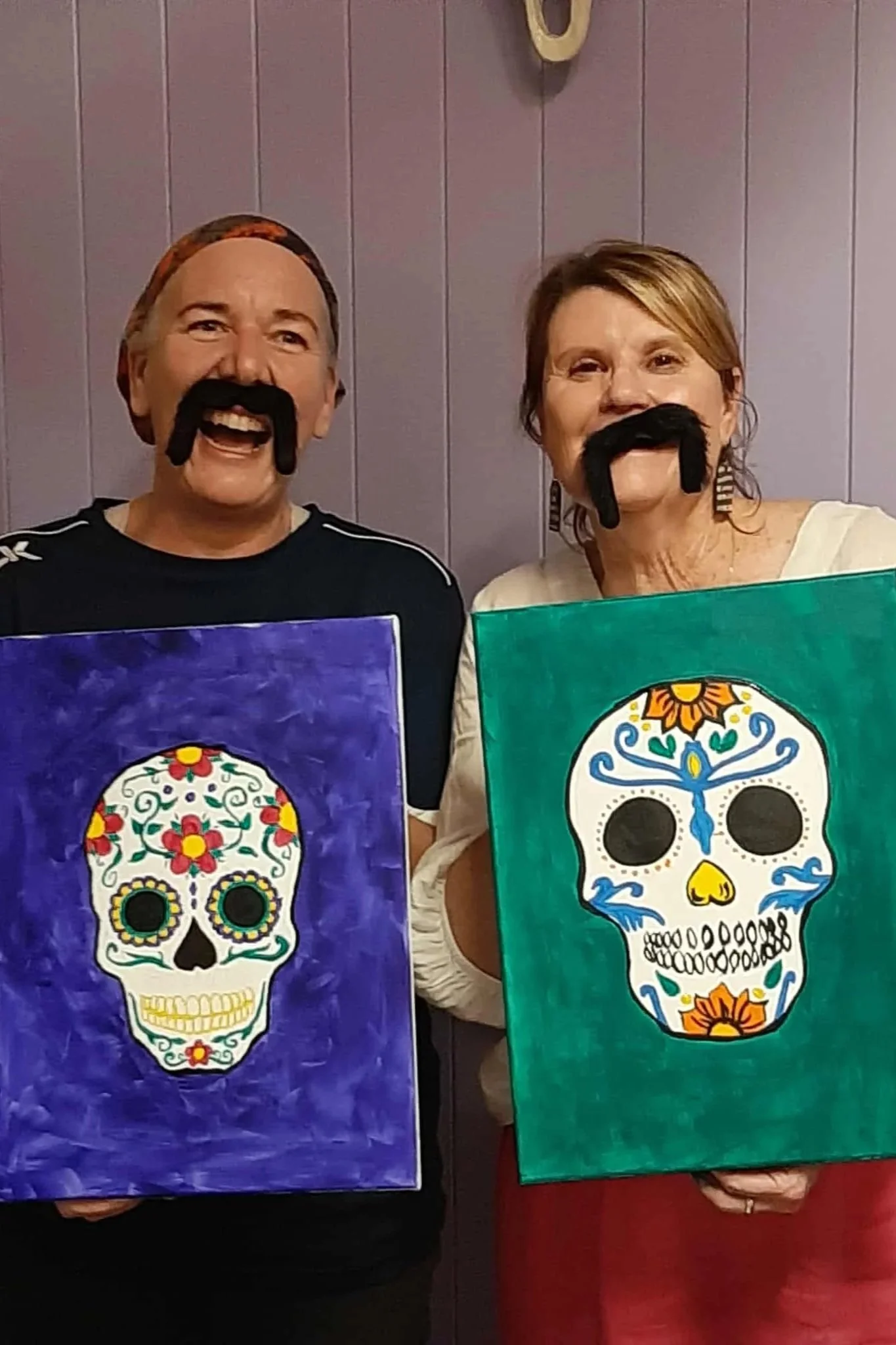 Two people smiling and wearing fake black mustaches, holding colorful paintings of decorated sugar skulls.