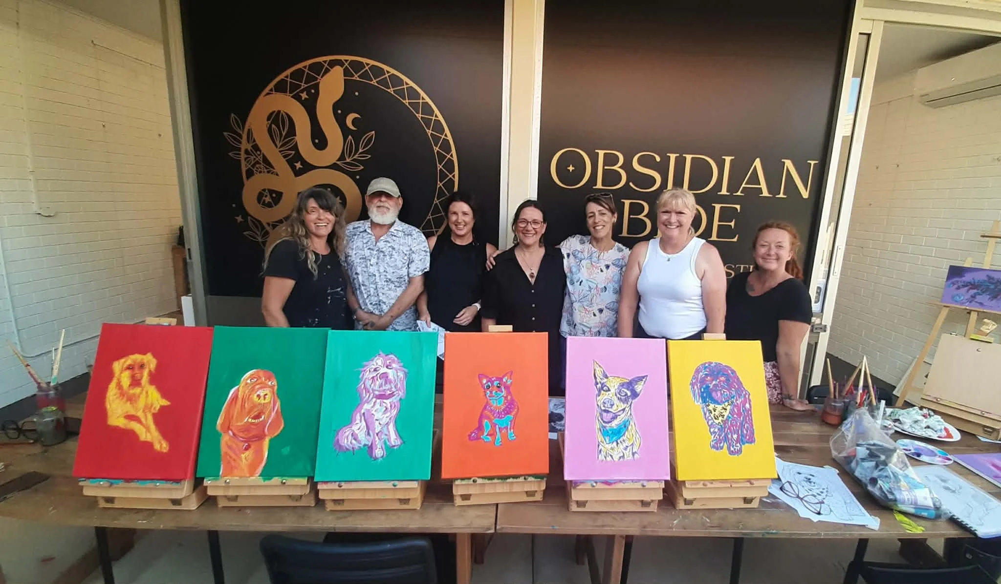 Group of seven people standing behind a table with colorful dog portrait paintings on display, inside an art studio with the sign 'Obsidian Babe Studio' in the background.
