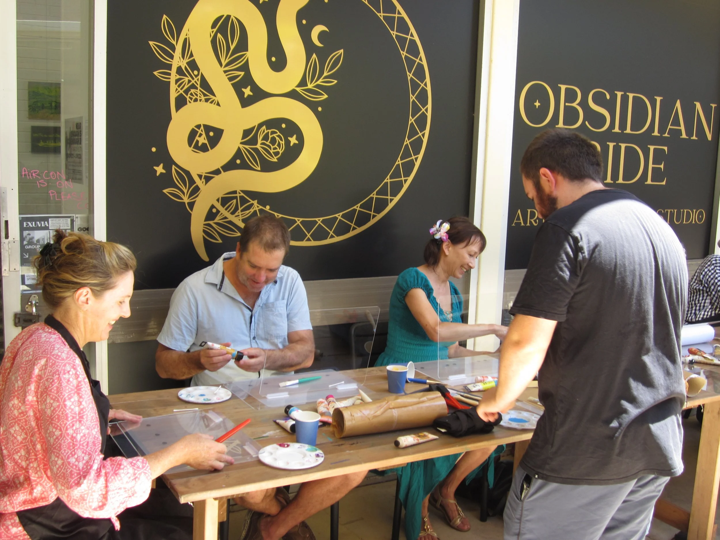 People participating in a jewelry-making workshop at a table, with an artisan assisting, in front of a black wall with gold art and the words 'Obsidian Guide' and 'Art Studio'.