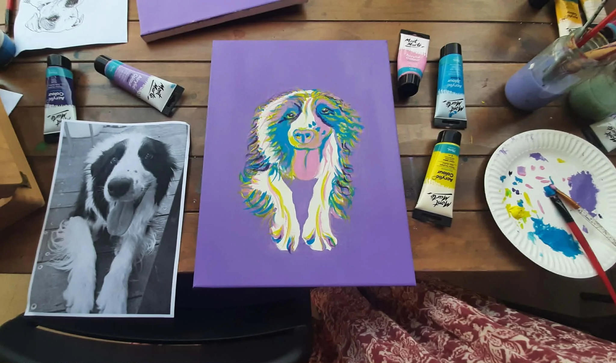 Colorful acrylic painting of a dog with long ears on purple background, surrounded by paint tubes, a black and white photo of a dog, and art supplies on wooden table.