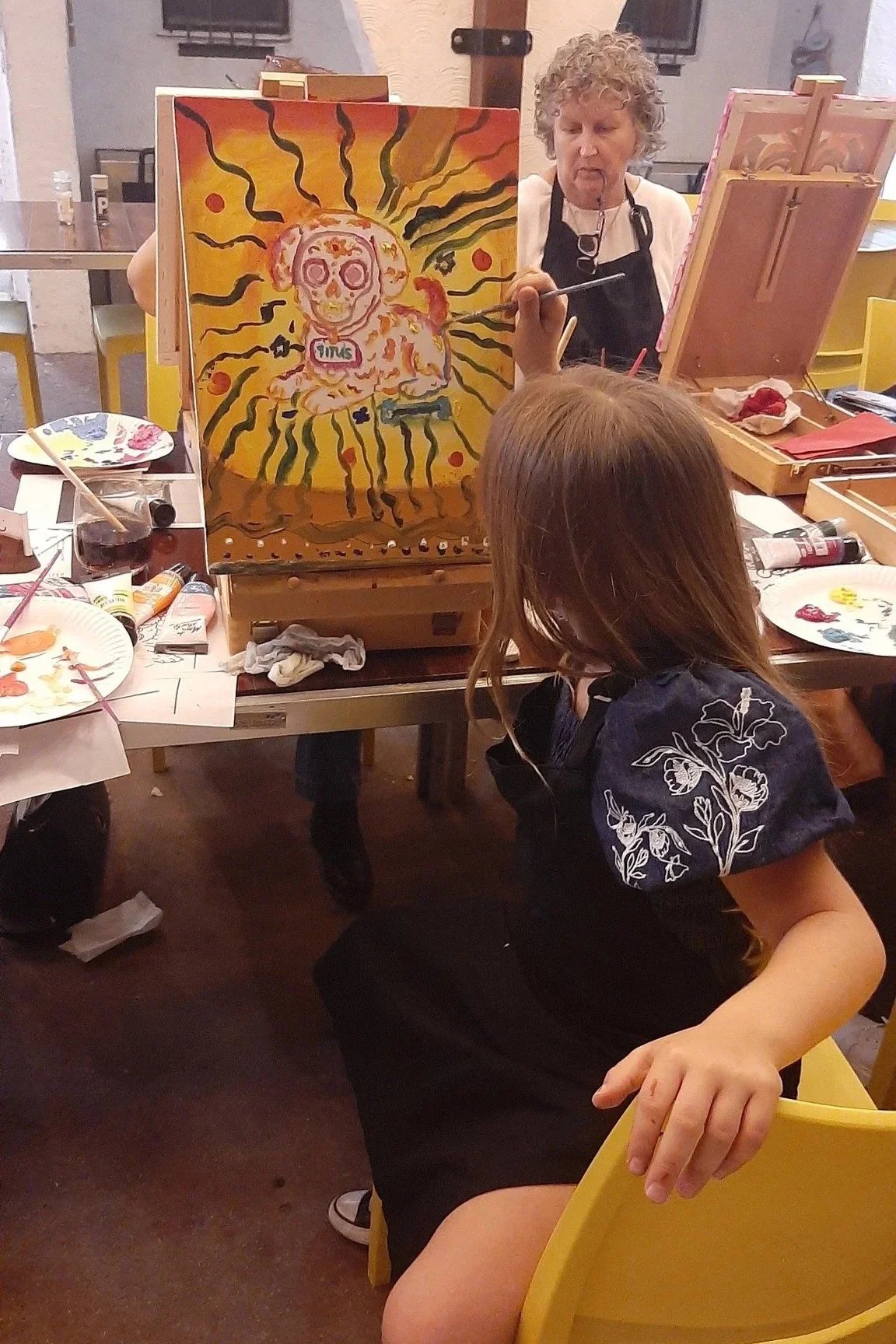 A woman is painting a colorful portrait of a skull with a flower crown on a canvas, while a girl watches in a studio setting. The painting features bright yellow, orange, pink, and black colors.