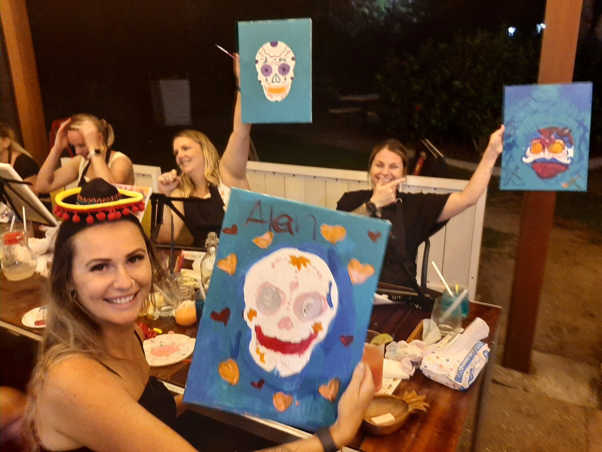 A group of women at an art party celebrating Dia de los Muertos. One woman in the foreground holds a colorful painting of a sugar skull surrounded by hearts. Some women are wearing hats and festive accessories, with paintings of sugar skulls on display and art supplies on the table.
