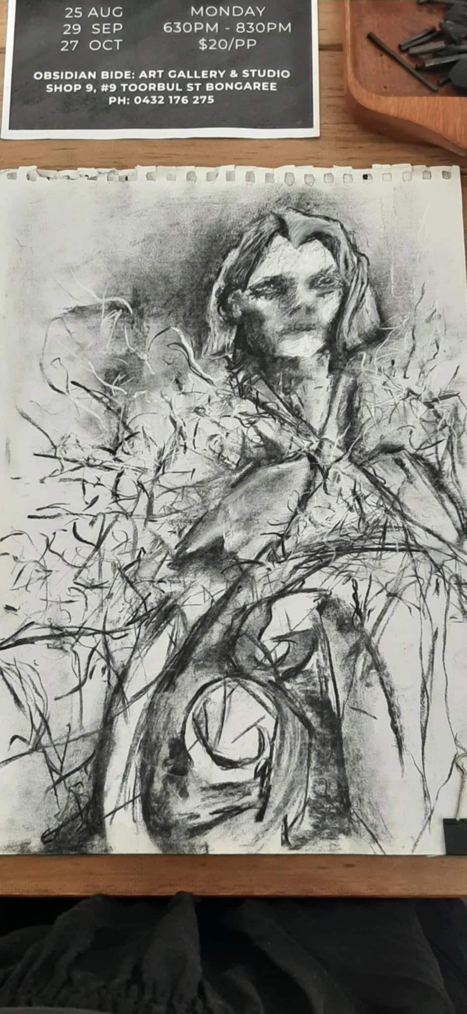 A charcoal sketch of a woman with hair parted in the middle, surrounded by abstract, tree-like lines with branches and leaves. There's a partially visible event poster at the top with details about an art event.