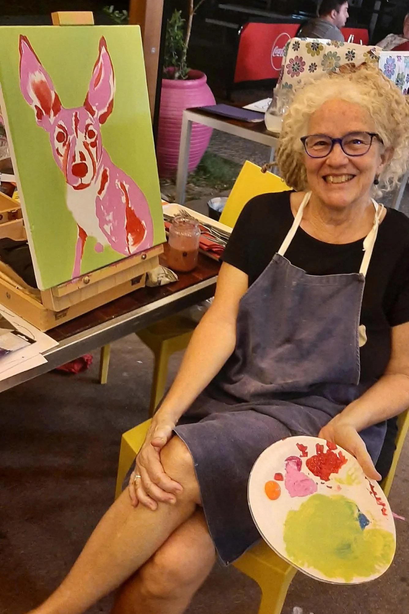 An elderly woman with curly blonde hair, glasses, and a black shirt sitting on a yellow chair, smiling at the camera. She is wearing an apron and holding a palette with paint. In front of her is a painting of a pink and red dog on a green background, created with bold brushstrokes.