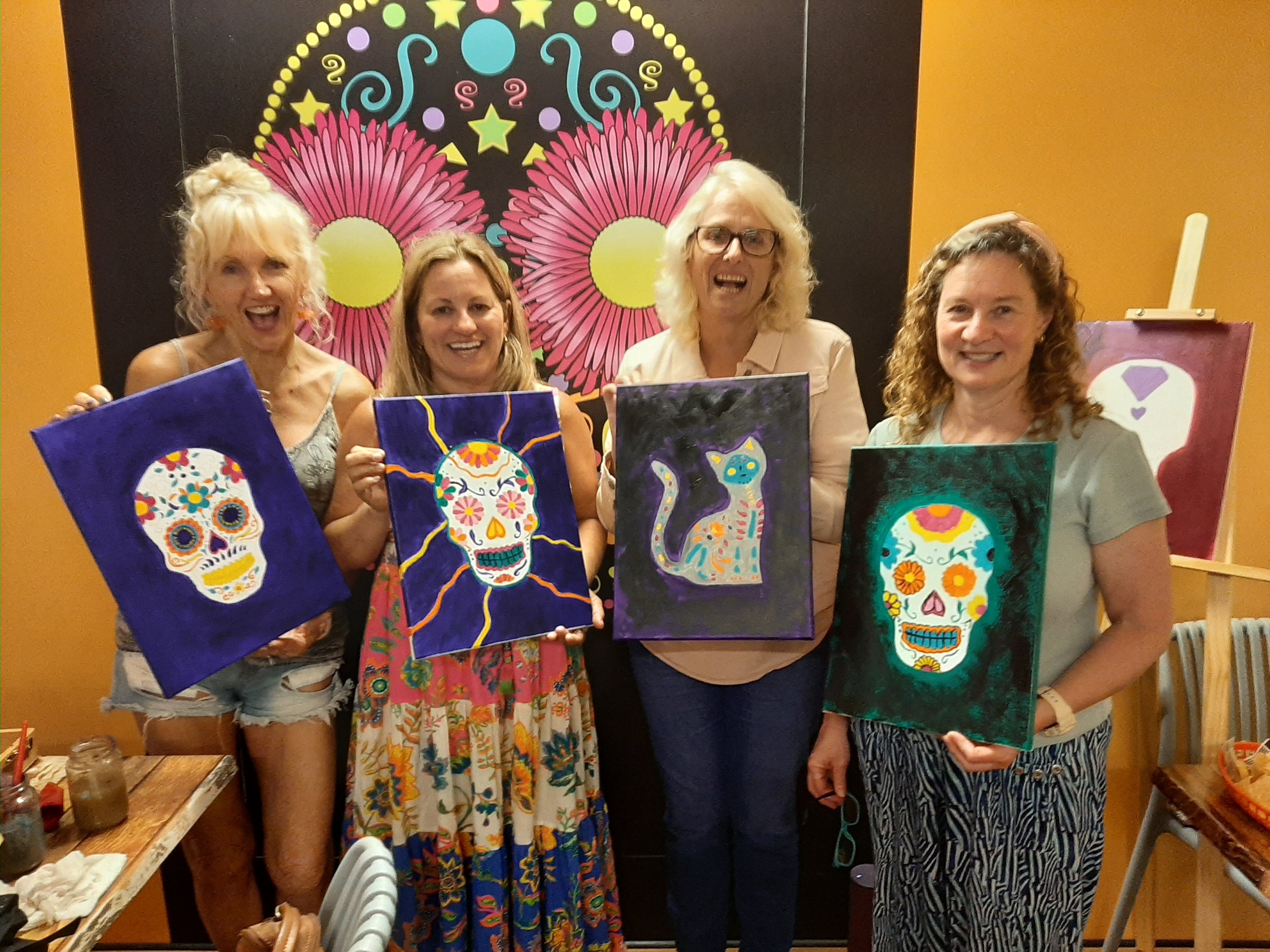 Five women standing in a colorful art studio, holding up paintings of decorated sugar skulls and a cat, with bright mural artwork of pink and yellow flowers behind them.