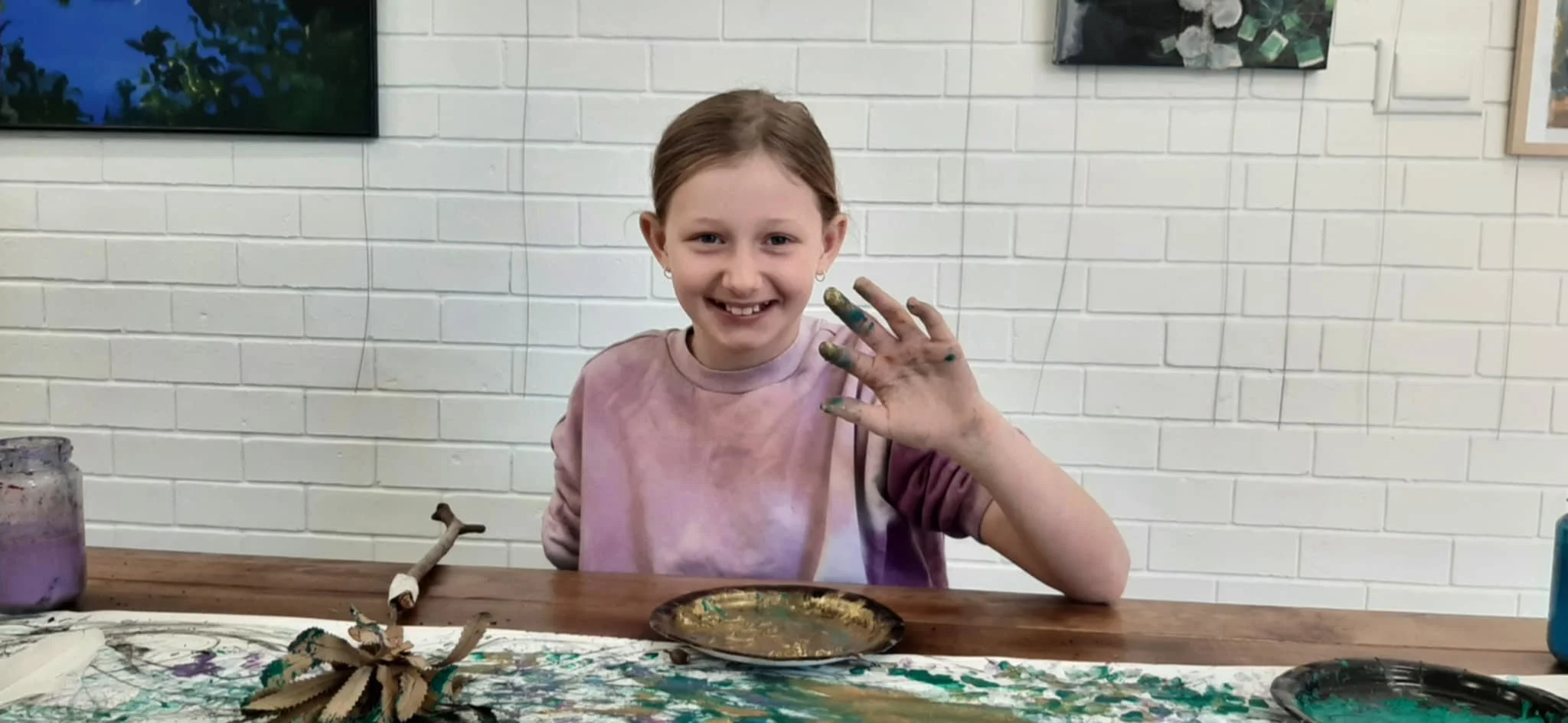 Young girl with paint on her hands smiling and showing her painted fingers at a table with art supplies, against a white brick wall.