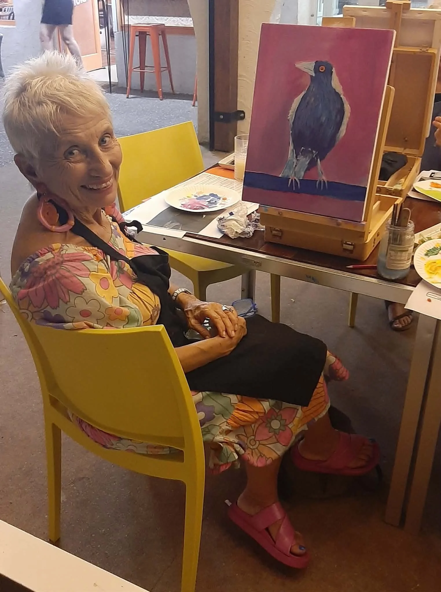 An elderly woman sitting at a table with a painting of a bird in progress on an easel in front of her, smiling at the camera. She is wearing a floral dress and pink sandals, with art supplies and a plate of paint on the table.