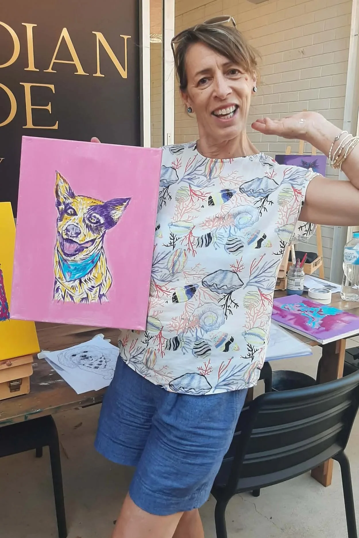 A woman smiling and holding a painting of a dog wearing a blue bandana, standing indoors at an art event.