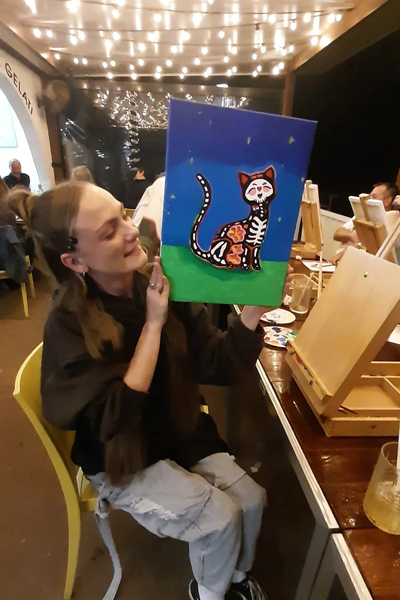 A woman sitting at a table holding up a colorful painting of a cat with floral patterns inside and a skeleton face, in a lively restaurant or event space with string lights overhead.