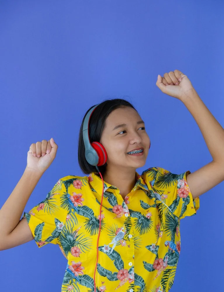 Young girl with a yellow floral shirt smiling and dancing while wearing red and gray headphones against a blue background for Sunnyside Homeschooling's StarJam Homeschool Music Program