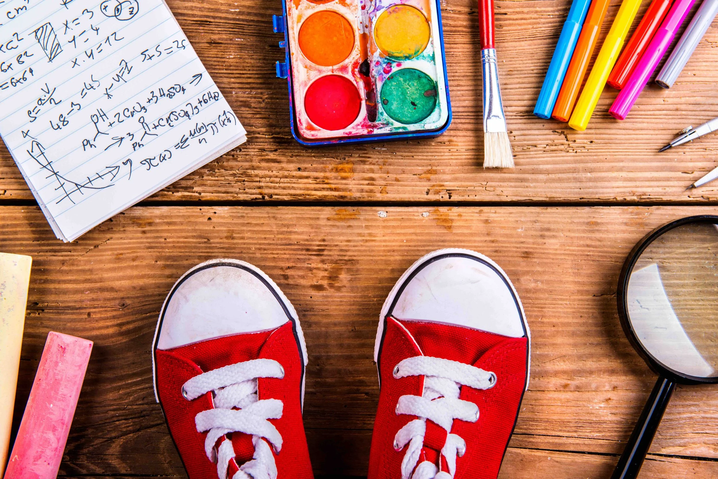 A person wearing red and white sneakers standing on a wooden floor, with a watercolor paint set, colored markers, a paintbrush, and a notebook with math notes nearby for Sunnyside Homeschooling's Programs