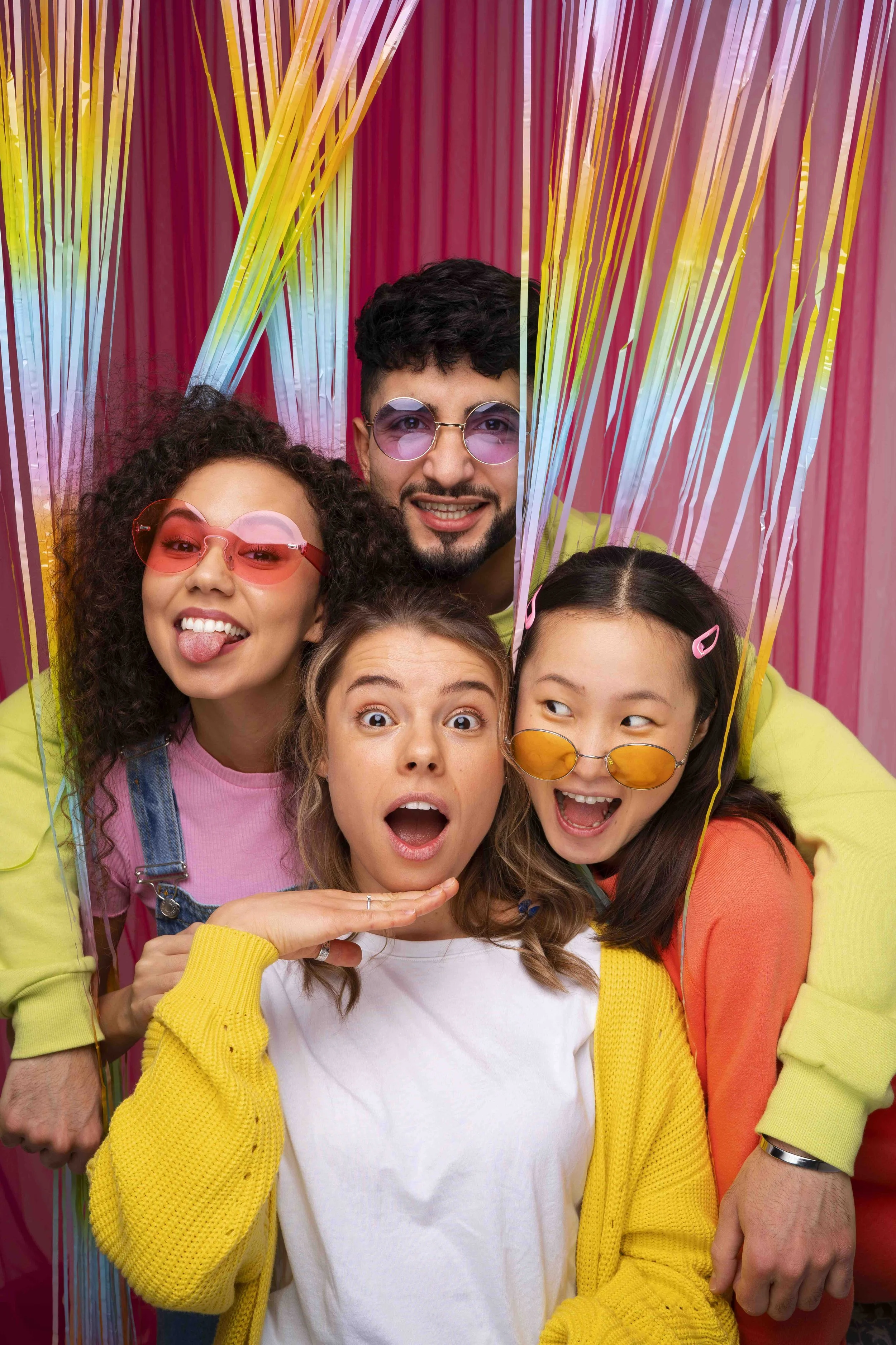 A group of five young friends posing together behind colorful, striped decorations. They are wearing vibrant clothing and sunglasses, making silly faces.