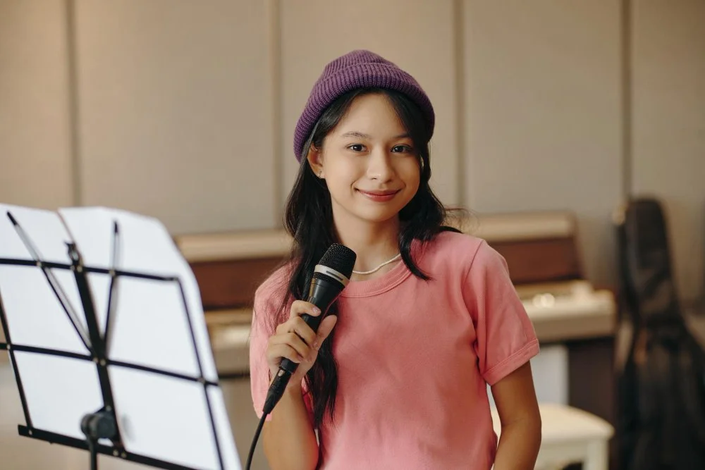 A young woman with long dark hair wearing a purple beanie and pink t-shirt holding a microphone, standing next to a music stand in the StarJam Homeschool music Program by SoundsKradle and Sunnyside Homeschooling Community
