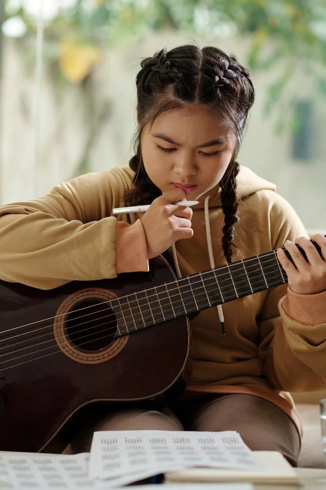 Young girl with braided hair thoughtfully practicing guitar and reading sheet music indoorsnfor Sunnyside Homeschooling's StarJam Homeschool Music Program.
