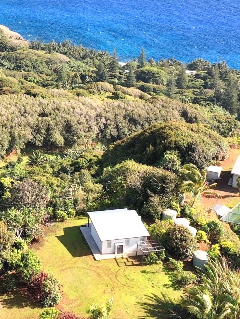 Aerial view of a small white house surrounded by green vegetation near a coastline with blue ocean in the background.