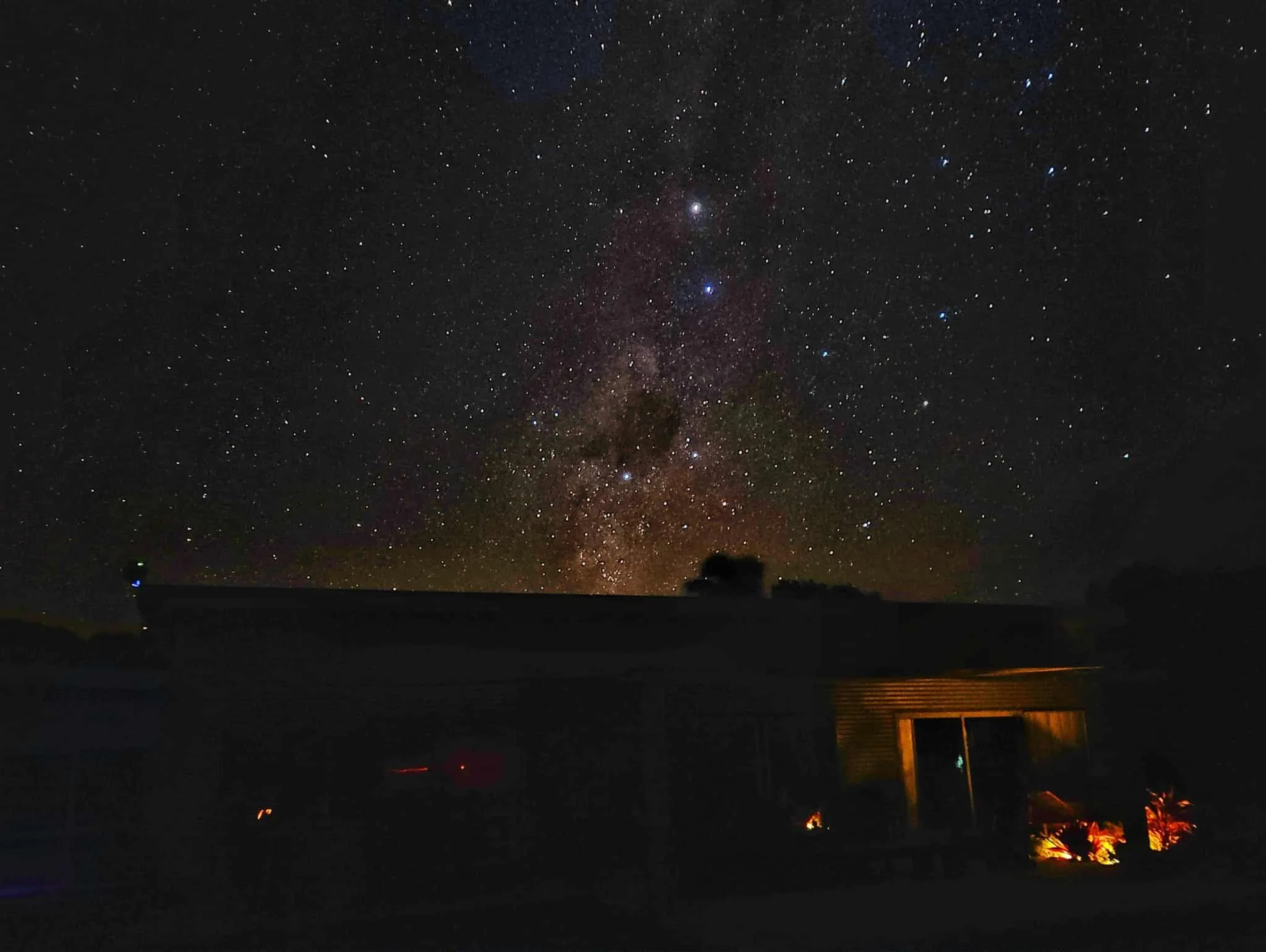 Night sky filled with stars and the Milky Way galaxy over a silhouette of a house with outdoor lighting.