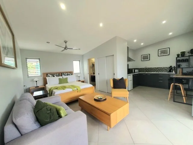 Modern studio apartment interior with a bed, sofa, coffee table, and kitchenette. The room is decorated with neutral colors, framed pictures, and a ceiling fan.